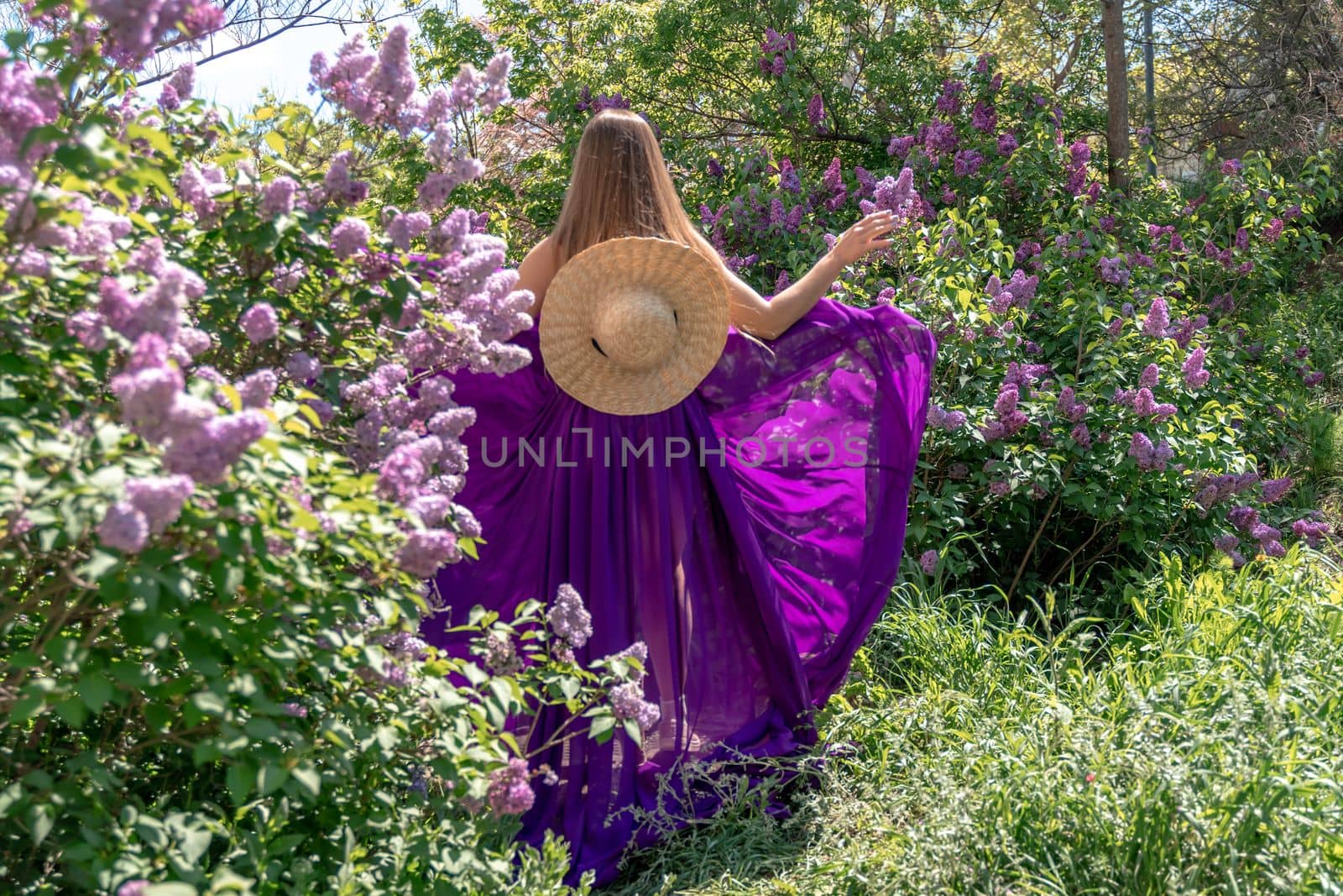 Fashion Model in Lilac Flowers, Young Woman in Beautiful Long Dress Waving on Wind, Outdoor Beauty Portrait in Blooming Garden by Matiunina