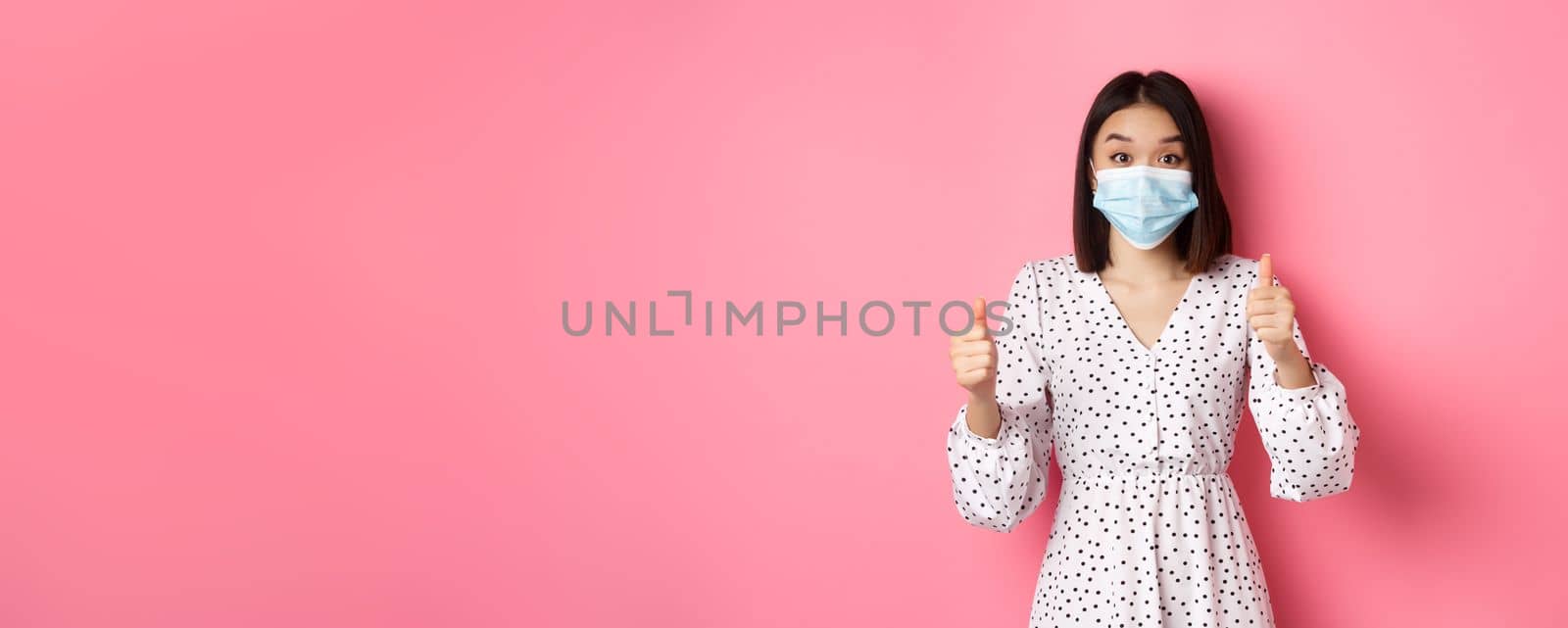 Covid-19, quarantine and lifestyle concept. Excited asian woman in face mask showing thumbs-up, praising good job, looking amazed at camera, standing over pink background by Benzoix