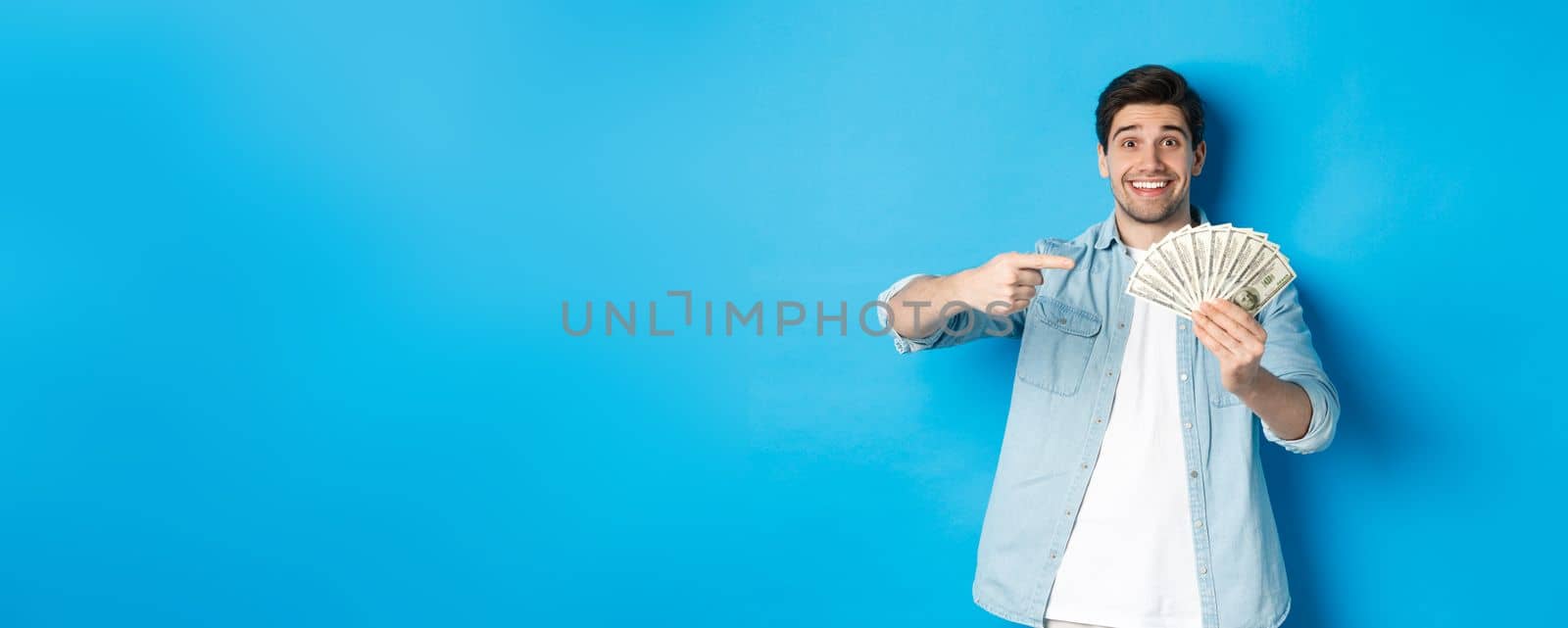 Surprised smiling man in casual clothes, pointing fingers at money, standing over blue background.