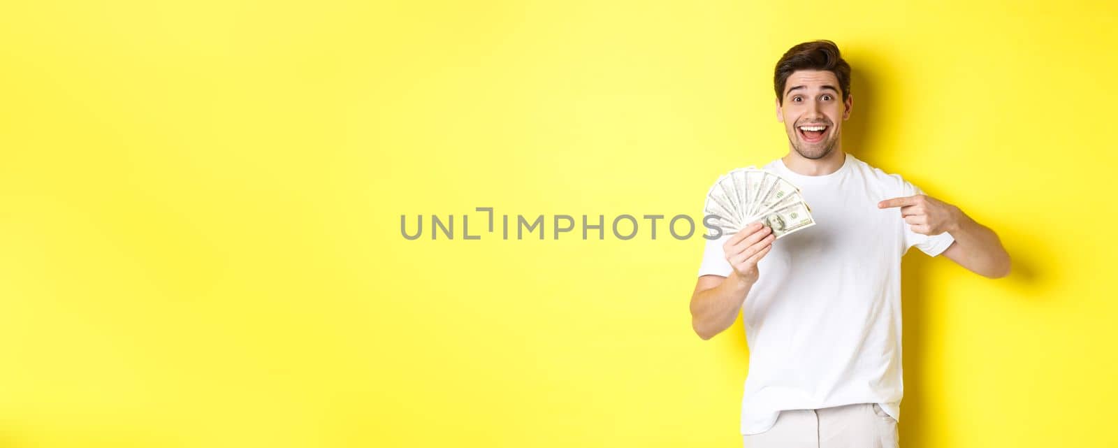 Happy handsome guy pointing finger at money, concept of credit and loan, standing over yellow background.