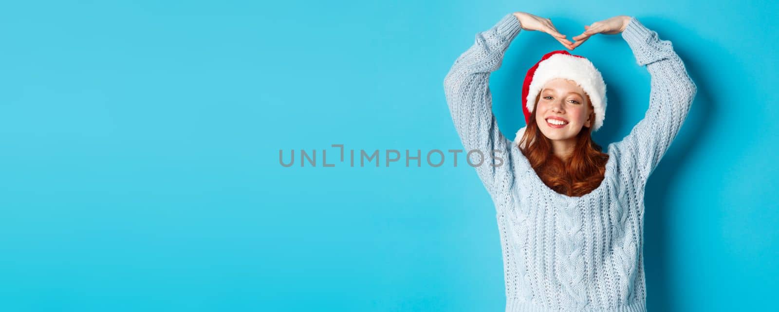 Winter holidays and Christmas Eve concept. Cute redhead teen girl in santa hat and sweater, making heart sign and smiling, wishing merry xmas, standing over blue background.