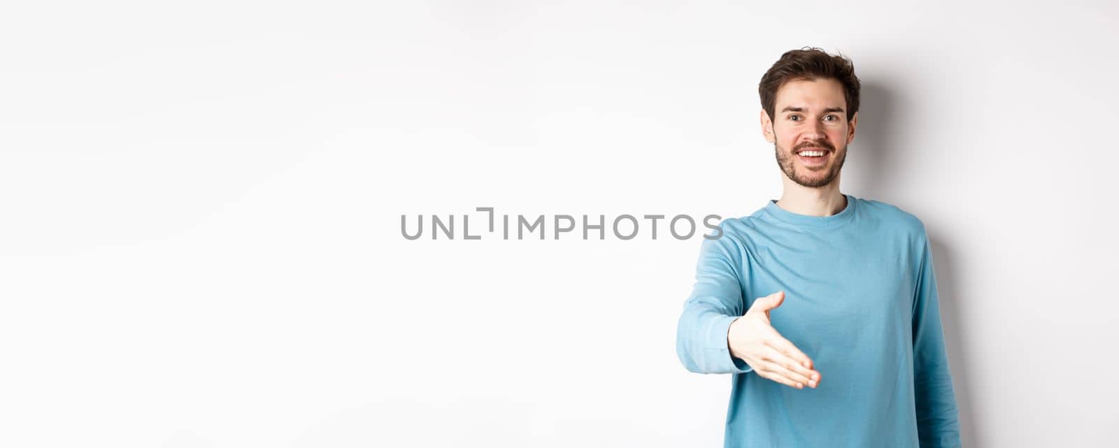Friendly smiling man stretch out hand, saying hello and give arm for handshake, standing over white background.