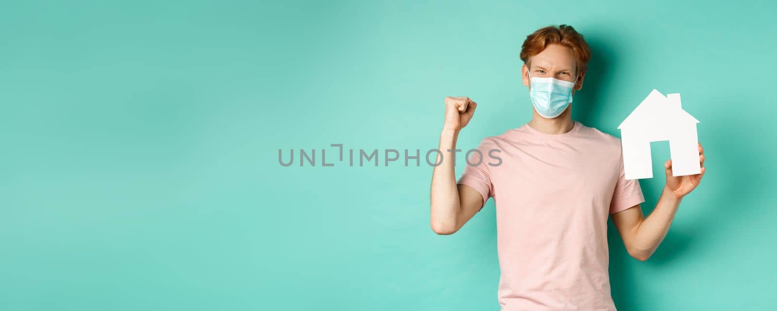 Covid-19 and real estate concept. Happy redhead man in medical mask, showing paper house cutout and fist pump, rejoicing and winning, standing over turquoise background by Benzoix