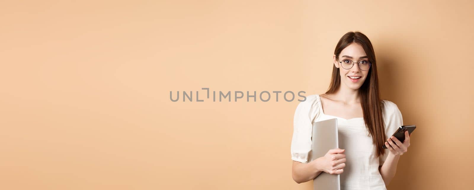 Smiling young woman in glasses reading exciting news on mobile phone, holding laptop and looking happy at camera, standing on beige background by Benzoix