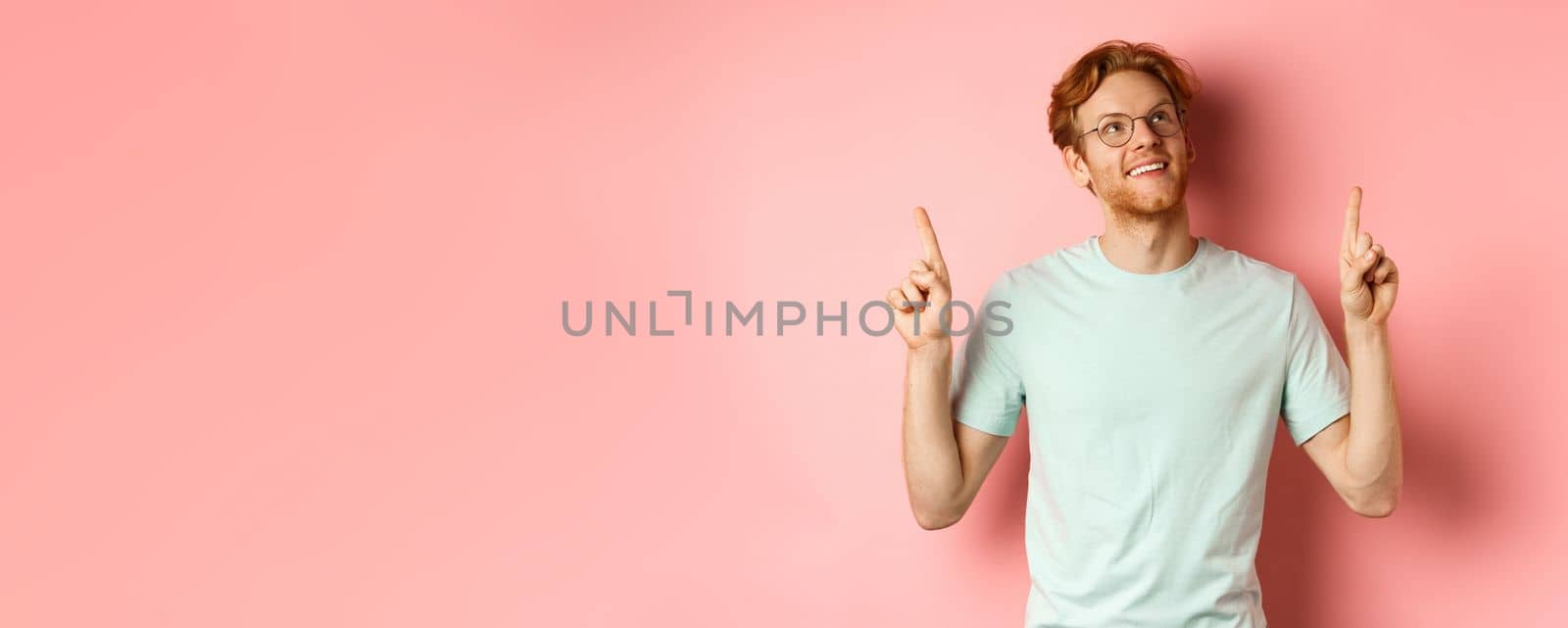 Dreamy young man with red hair and beard pointing fingers and looking cheerful up, admire something, standing over pink background.