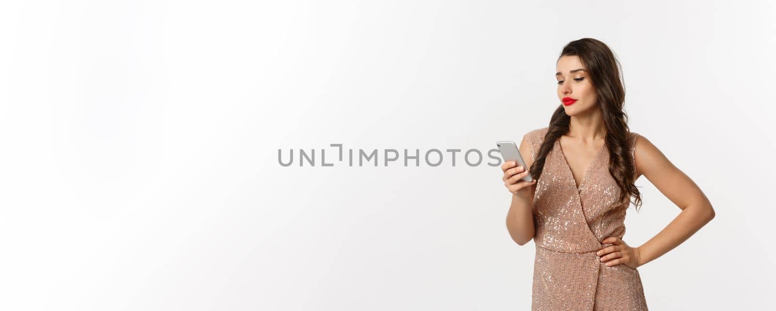 Christmas party and celebration concept. Careless woman in stylish dress reading message on mobile phone, looking skeptical, standing over white background.