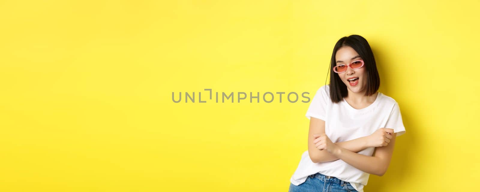 Fashion and lifestyle concept. Sassy and confident asian woman in trendy sunglasses looking self-assured at camera, standing over yellow background.