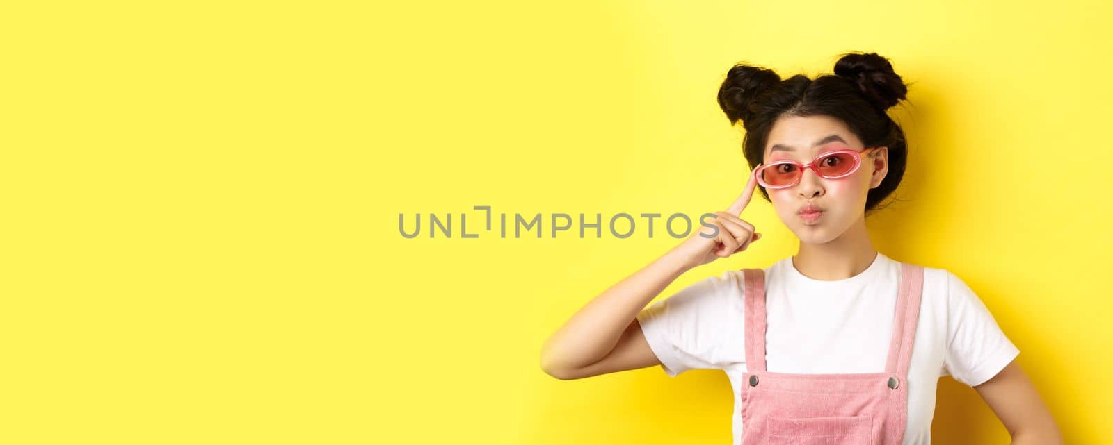 Stylish teen asian girl wearing sunglasses and pink overalls, standing on yellow background by Benzoix