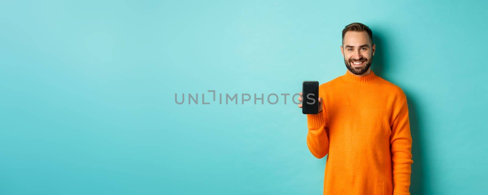 Handsome bearded guy in orange sweater, showing smartphone screen and smiling, showing promo online, turquoise background.