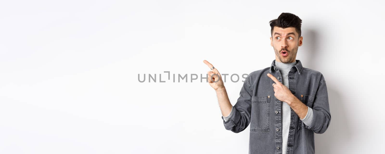 Stylish guy say wow, pointing and looking left with excited face, showing banner logo, standing on white background.