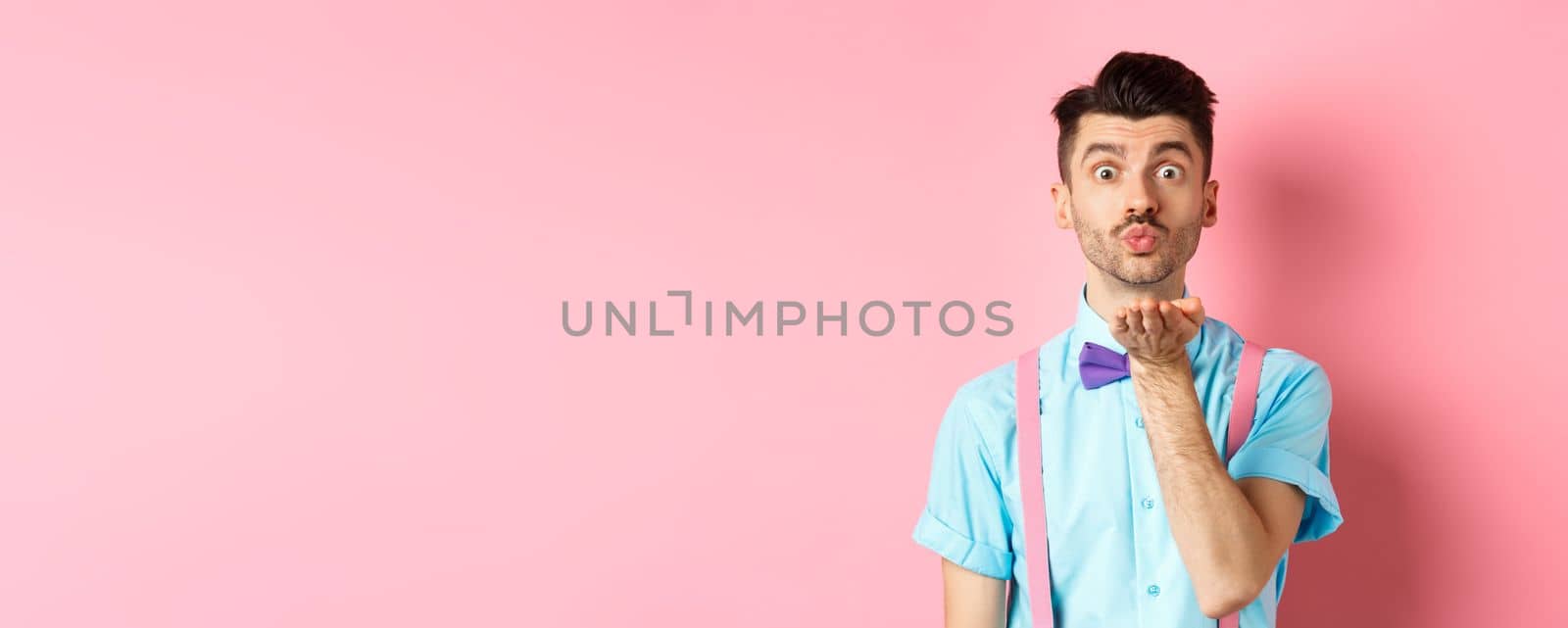 Cute boy sending air kiss at camera and saying I love you on Valentines day, standing over romantic pink background in bow-tie and shirt.