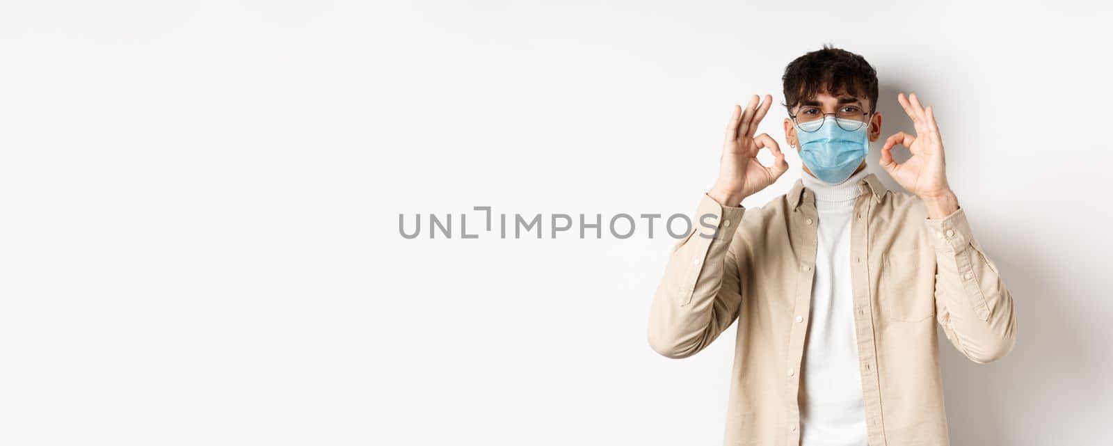 Covid-19, health and real people concept. Handsome guy in glasses and medical mask showing okay gesture in approval, recommend use preventive measures from corona, white background.