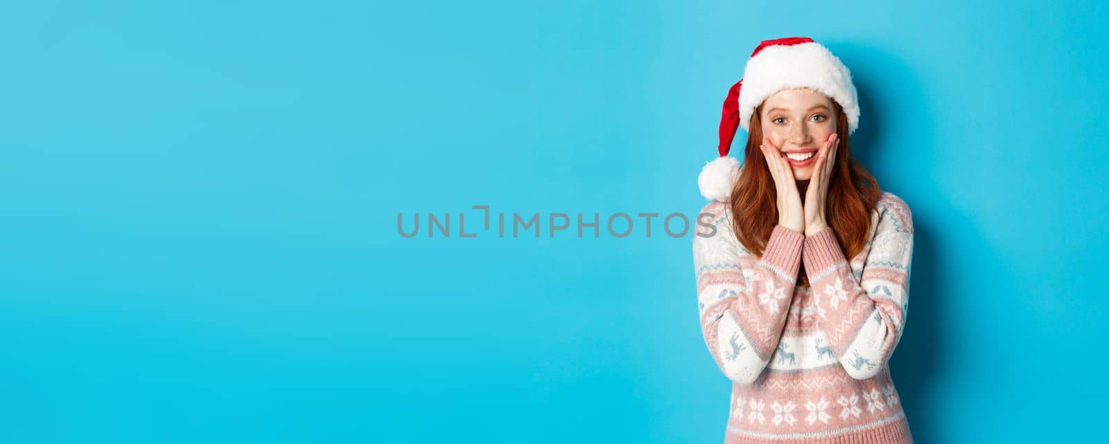 Winter and Christmas Eve concept. Happy redhead girl celebrating xmas, staring at camera amazed, smiling and touching cheeks, standing over blue background.