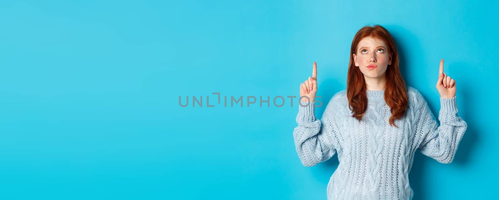 Winter holidays and people concept. Thoughtful redhead girl in sweater staring and pointing fingers up, having doubts, thinking or making choice, standing over blue background.