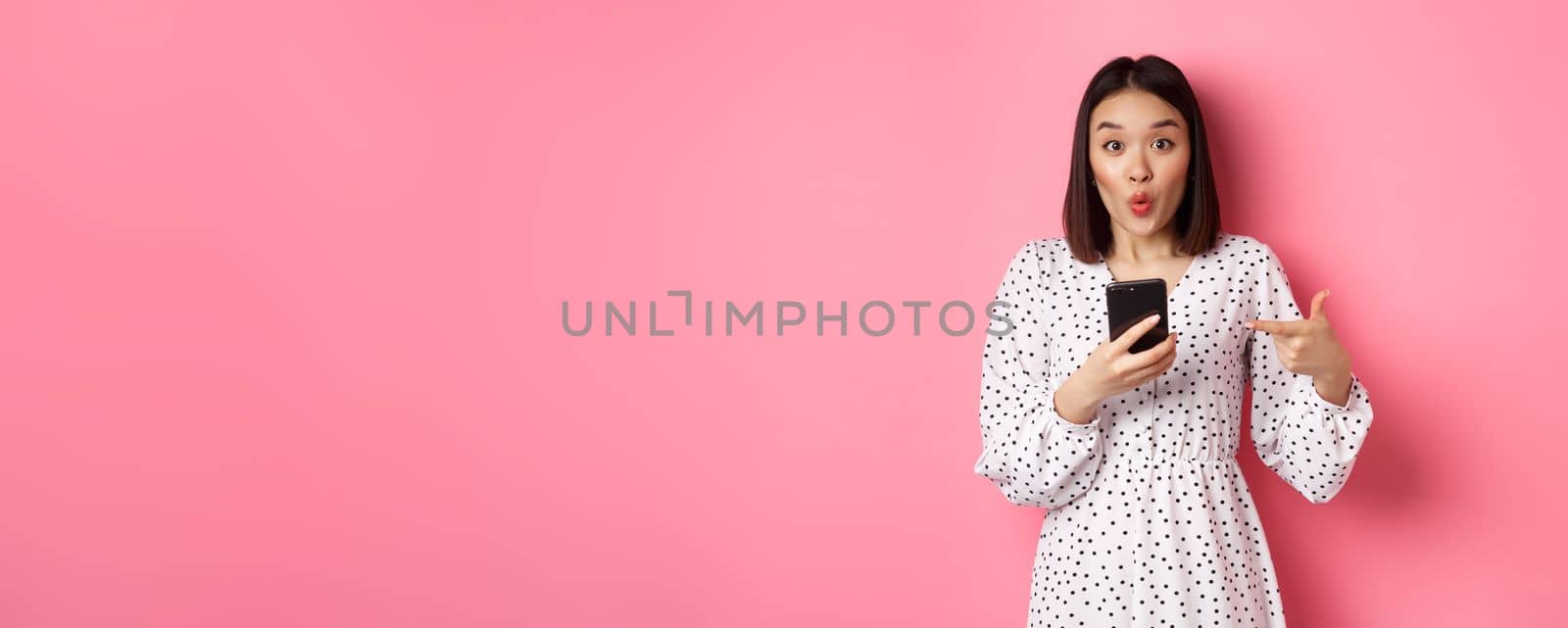 Online shopping and beauty concept. Amazed asian woman pointing finger at mobile phone, recommending smartphone app, standing over pink background.