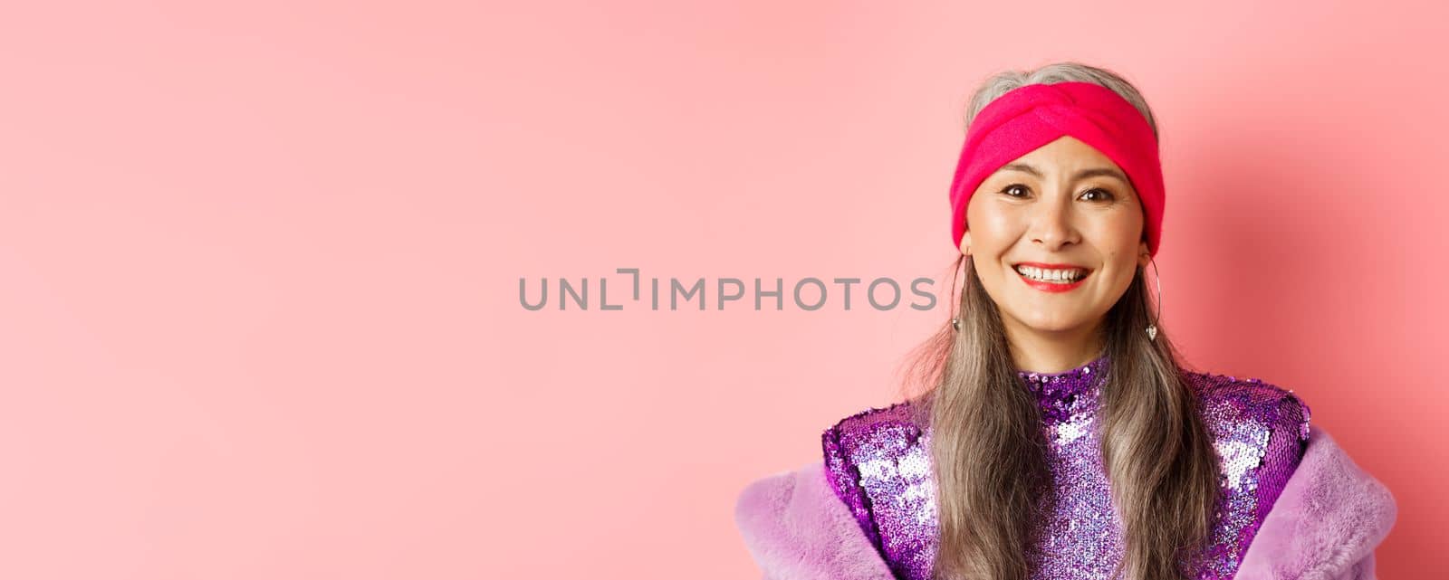 Close-up of trendy asian senior woman with grey hair and stylish disco dress, smiling happy at camera, standing over pink background.