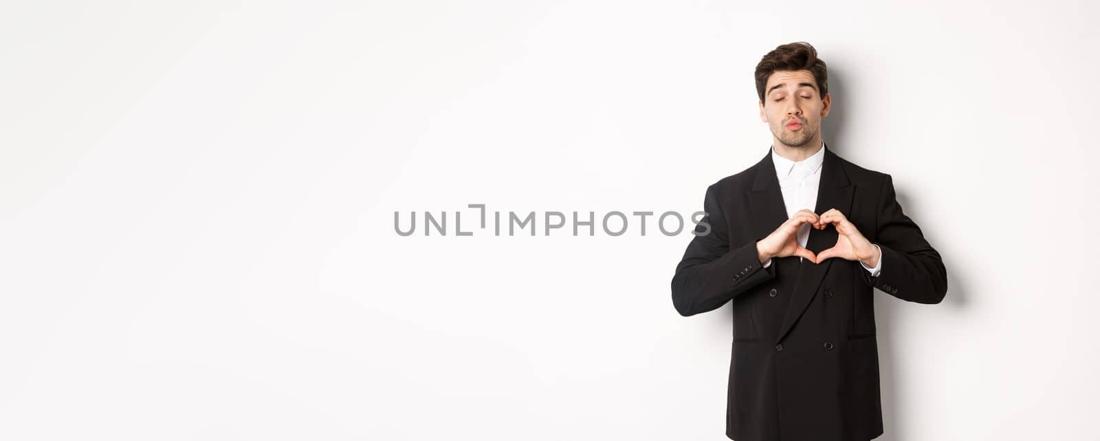 Image of handsome groom in black suit, showing heart sign, close eyes and pucker lips, waiting for kiss, standing against white background.