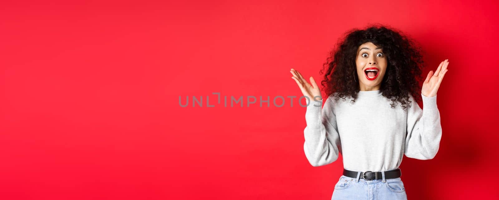 Image of happy and surprised curly woman in makeup and sweatshirt, raising hands up and rejoicing from good news, standing on red background.