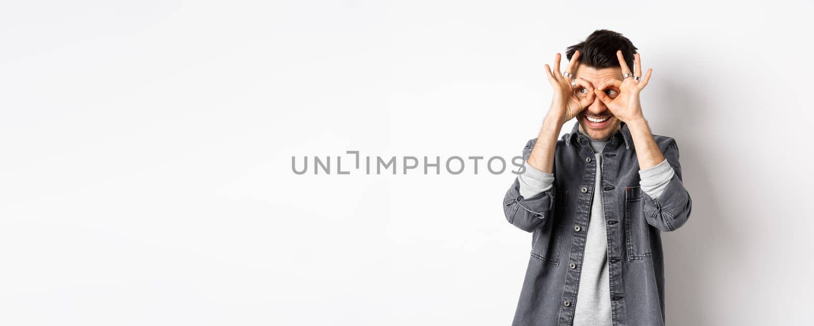 Smiling guy looking left at logo through hand binoculars, see good dea and look satisfied, standing on white background.
