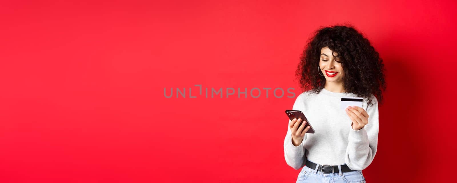 E-commerce and online shopping concept. Attractive caucasian woman paying for purchase in internet, holding smartphone and credit card, red background.