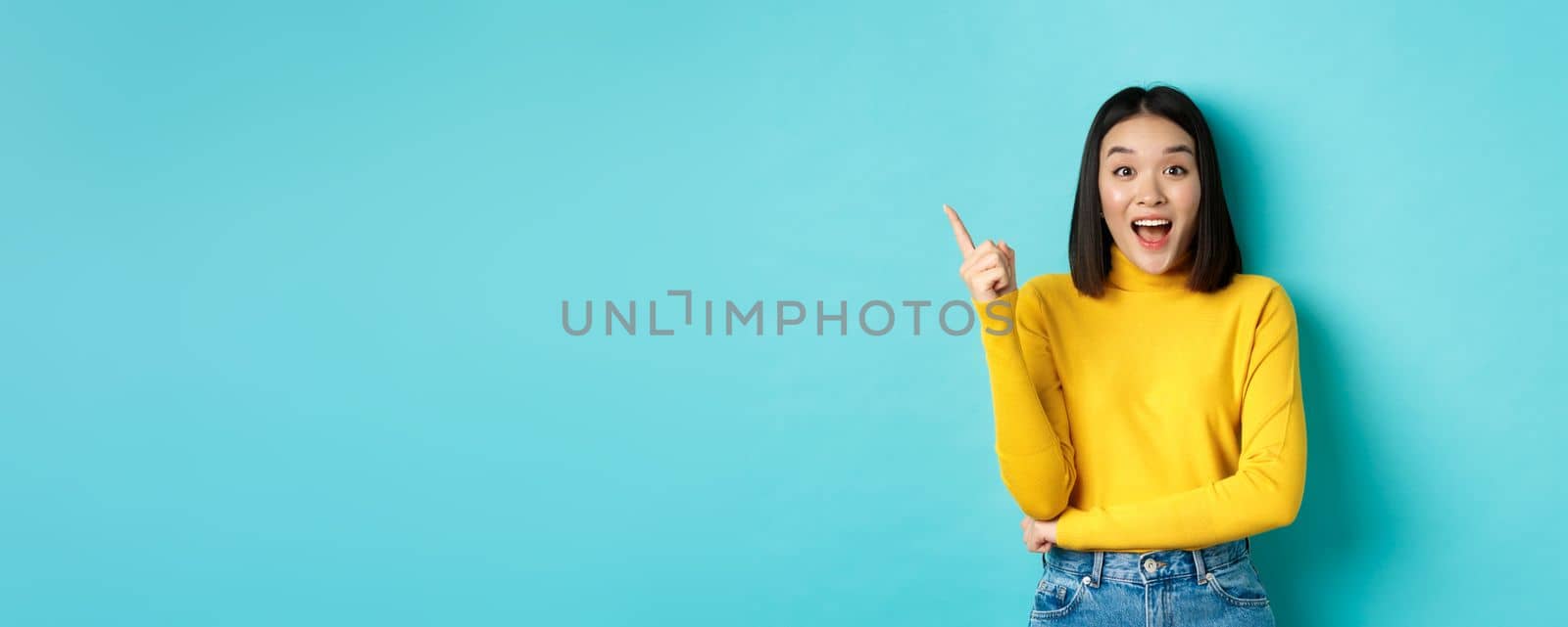Shopping concept. Attractive korean woman smiling amazed, pointing finger left, showing good deal banner, standing against blue background by Benzoix