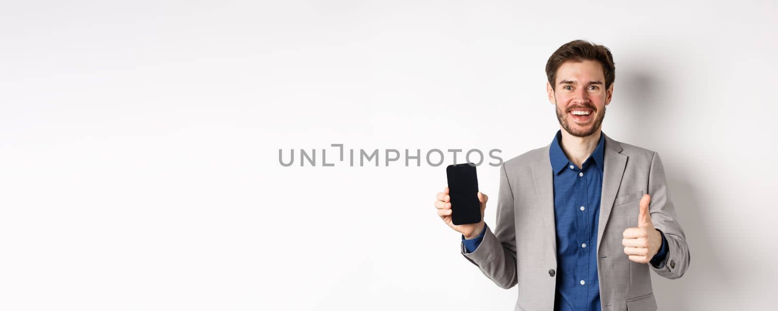 E-commerce and online shopping concept. Satisfied businessman in suit showing empty smartphone screen and thumb-up, praise good deal, white background.