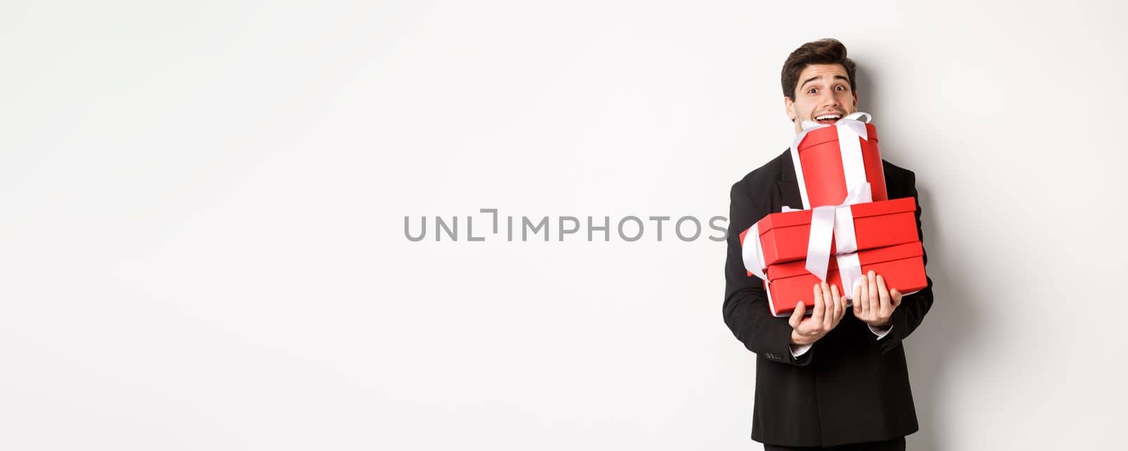 Concept of christmas holidays, celebration and lifestyle. Image of happy man in suit carry presents for new year, holding boxes with gifts and smiling, standing against white background by Benzoix