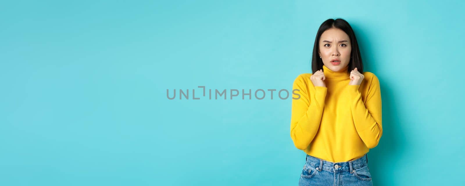 Image of worried asian woman with short dark hair, clench hands and stare at camera concerned, standing over blue background by Benzoix
