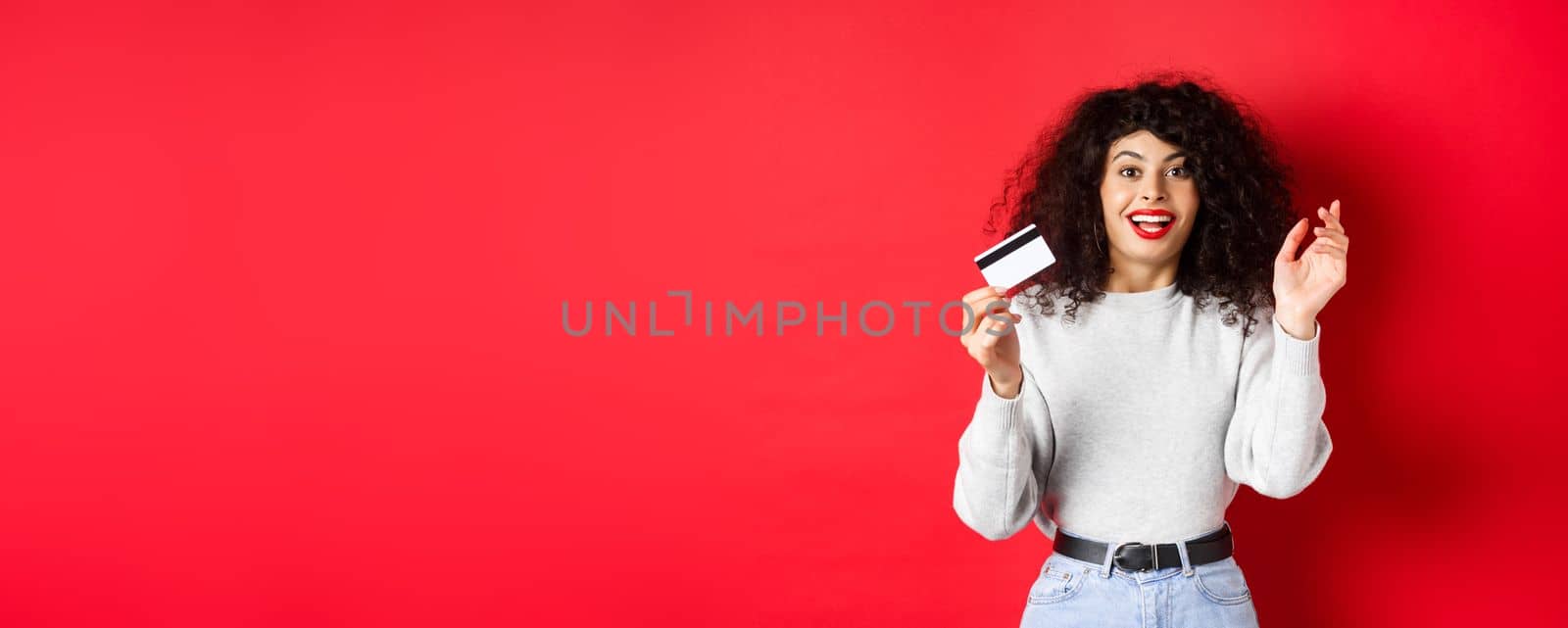 Excited woman telling about special deal, showing plastic card and looking happy, going shopping, standing on red background.