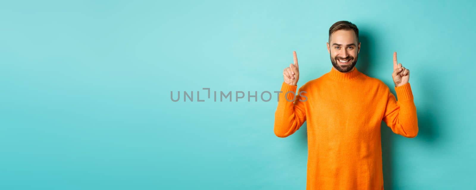 Handsome bearded guy in orange sweater showing up promo, pointing at top and smiling, standing over turquoise background.