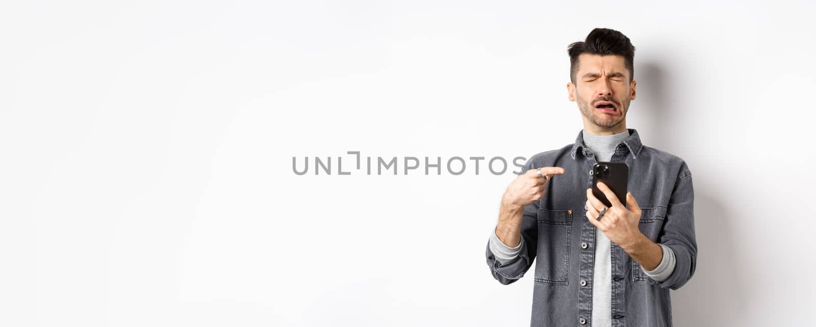 Sad crying guy pointing at smartphone and sobbing, complaining or feeling miserable, standing on white background.