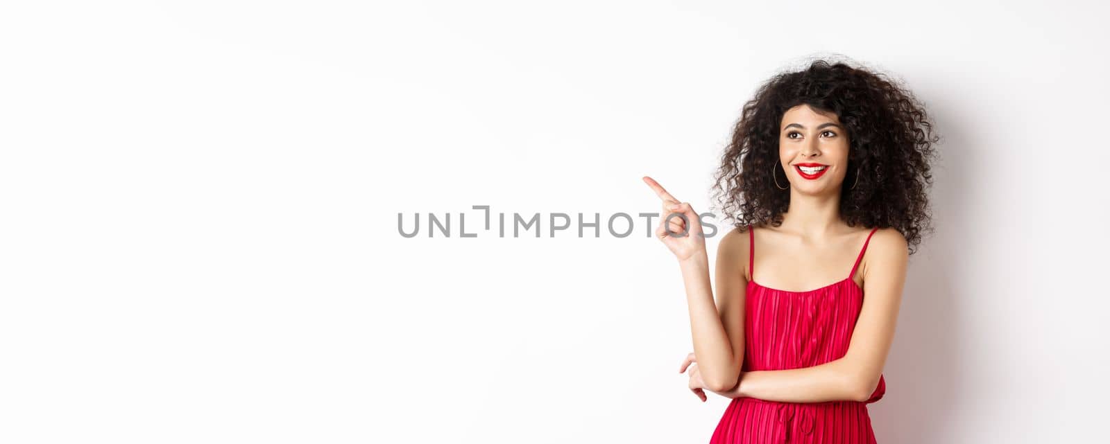 Elegant girl in red dress and evening makeup, pointing finger left and looking at logo, smiling satisfied, standing on white background.