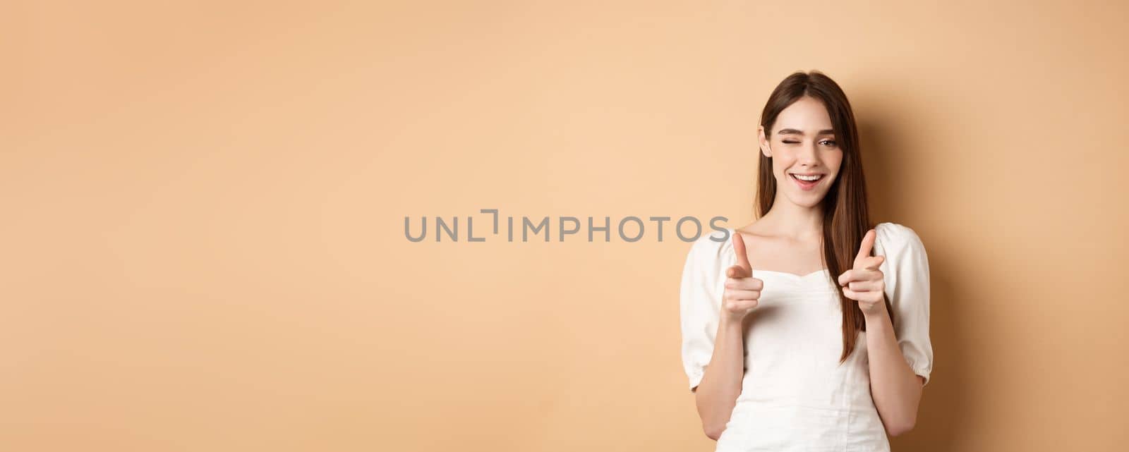 We need you. Smiling young woman pointing fingers at camera to beckon or invite people, standing on beige background.