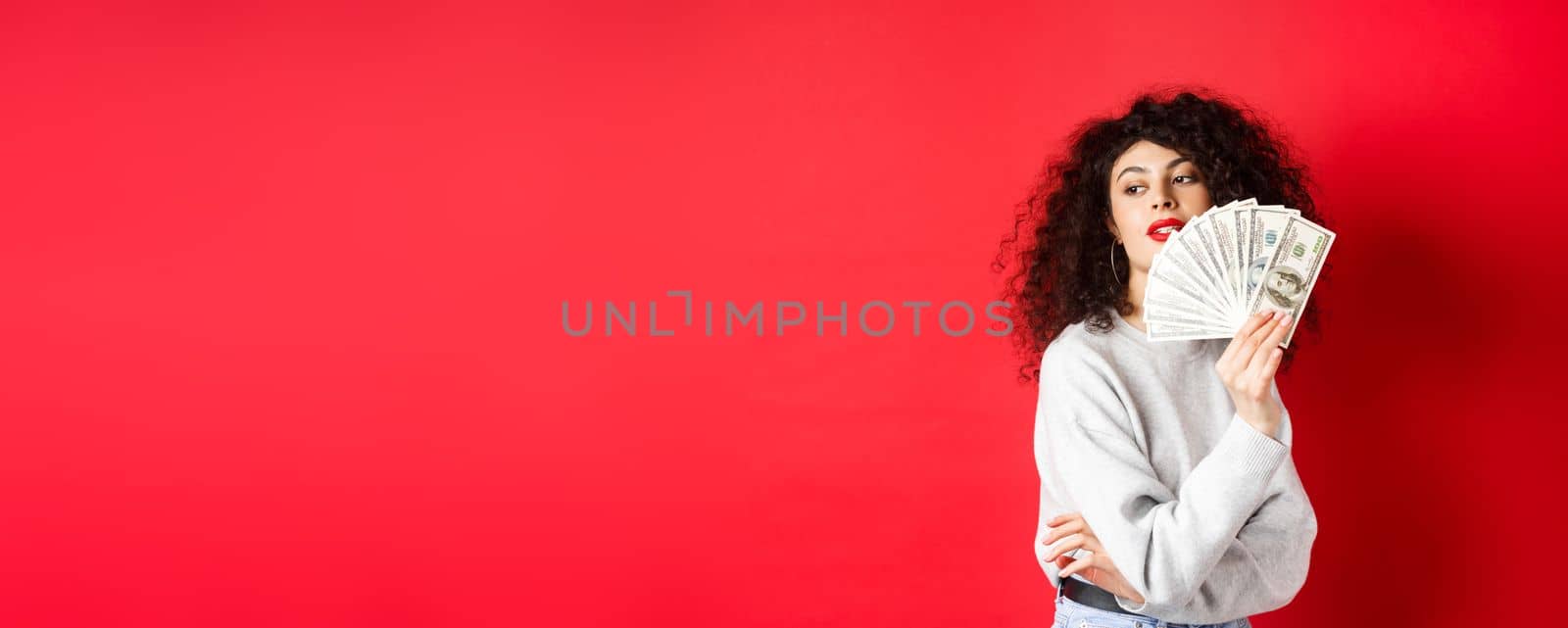 Beautiful rich woman looking sensual aside, waving at herself with dollar bills fan, standing seductive on red background by Benzoix