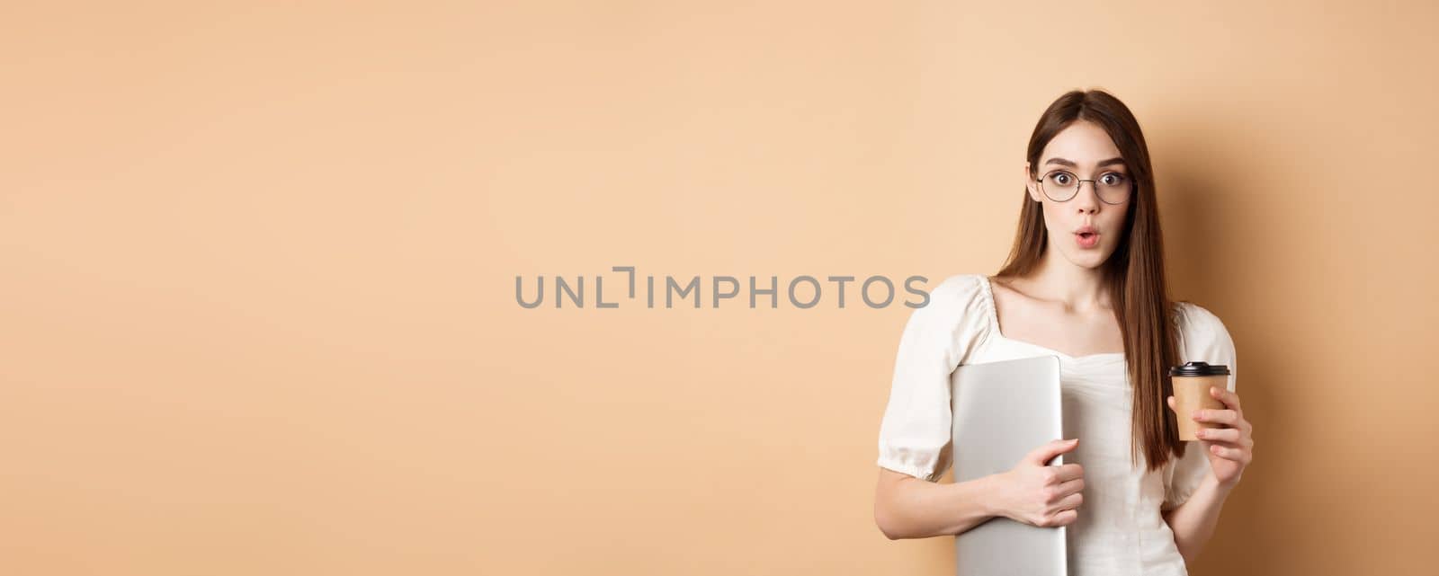 Working woman in glasses holding laptop and coffee, looking excited, hear amazing news, standing on beige background.