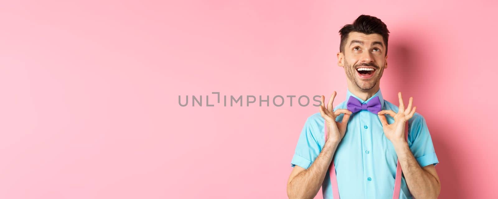 Happy young man with moustache, touching his bow-tie and laughing, looking up at logo, standing on pink background by Benzoix