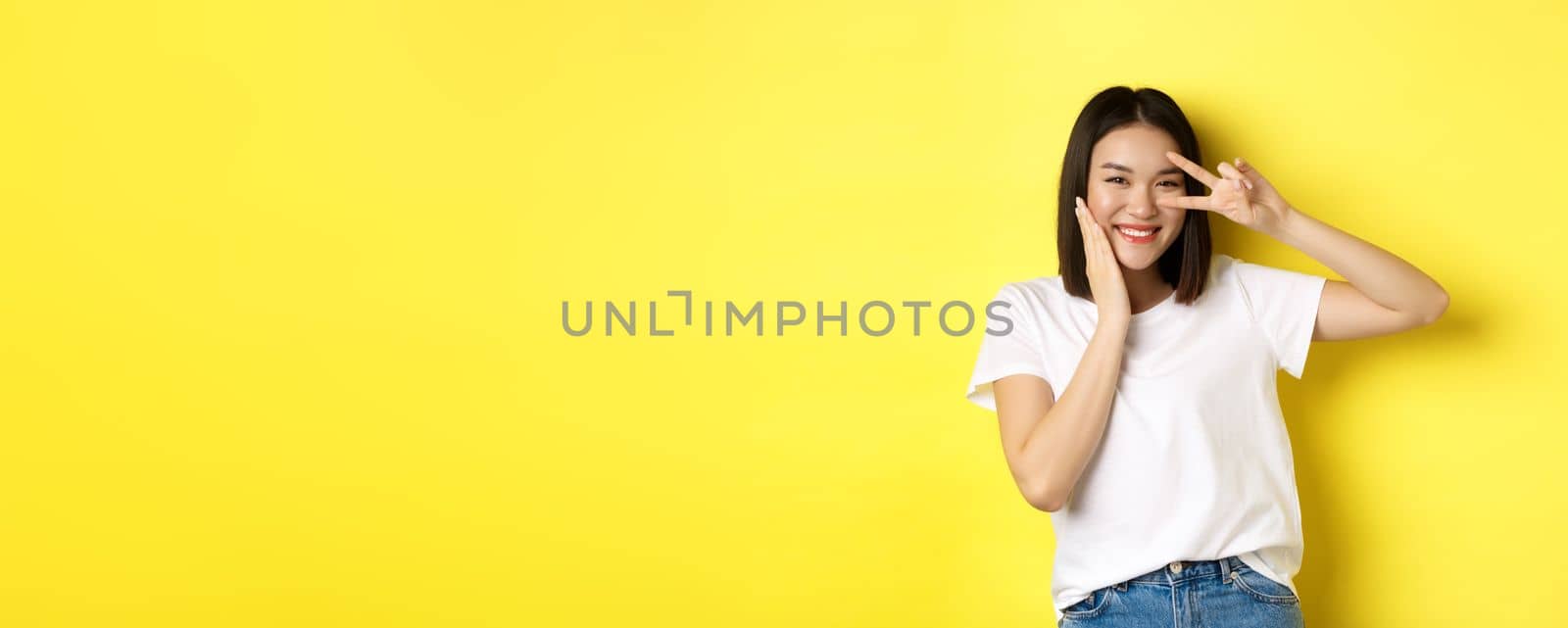 Lovely asian girl in white t-shirt posing with hand on cheek, showing peace sign on eye, standing over yellow background by Benzoix