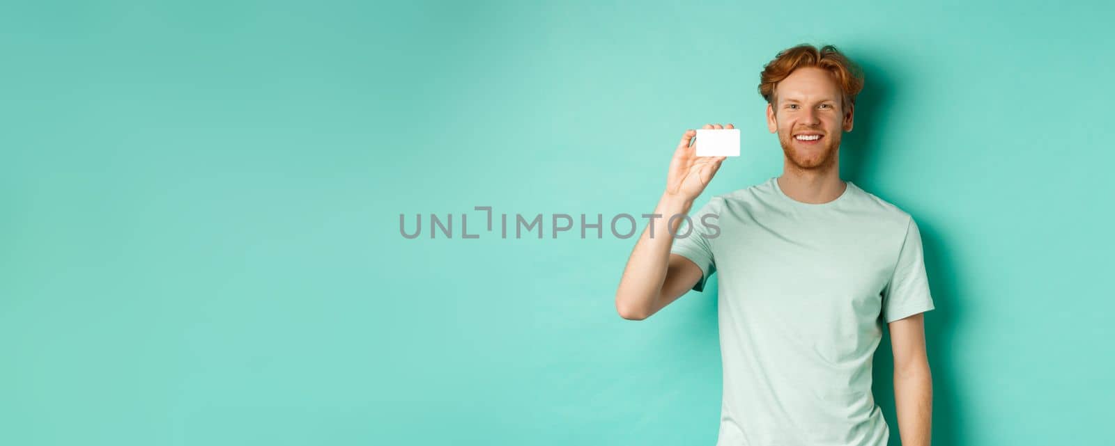Shopping concept. Handsome redhead man in t-shirt showing plastic credit card and smiling, standing over turquoise background by Benzoix