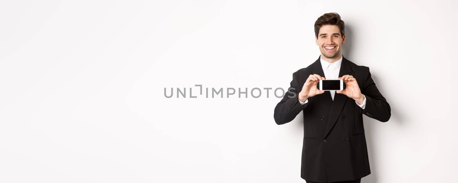 Portrait of attractive businessman in black suit, holding smartphone horizontally and showing screen, smiling pleased, standing against white background by Benzoix