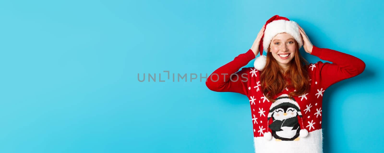 Happy holidays and Christmas concept. Pretty redhead girl in xmas sweater, put on santa hat and smiling, celebrating New Year, blue background.
