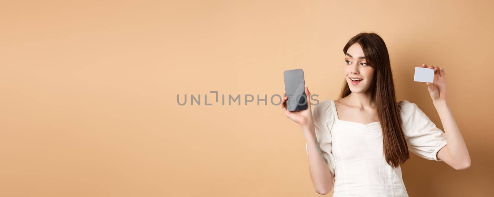 Check this out. Smiling happy young woman showing plastic credit card and empty mobile phone screen, demonstrate application, standing on beige background.