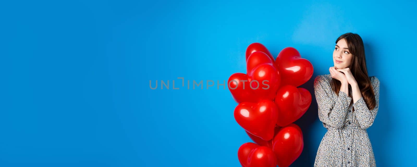 Valentines day. Dreamy beautiful woman in dress, imaging romantic date, standing near red hearts balloons and smiling, standing on blue background.