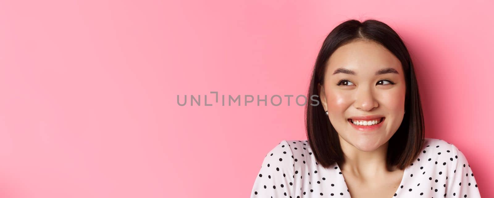 Beauty and lifestyle concept. Headshot of excited asian girl smiling, looking left at promo banner, standing over pink background. Copy space