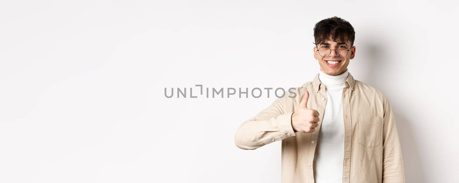 Positive smiling man showing thumbs up and looking satisfied, recommending and approving thing, standing pleased on white background.