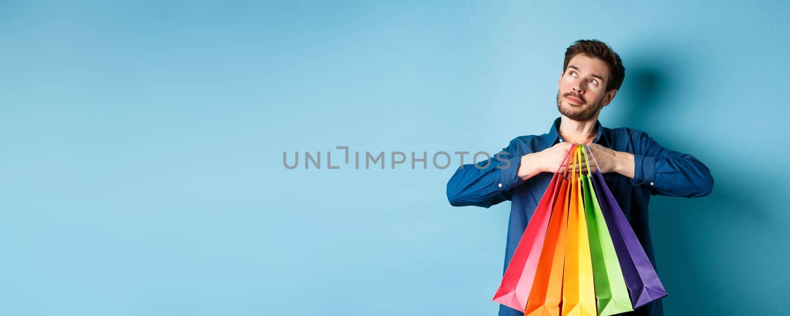 Dreamy young man holding colorful shopping bags and imaging something, looking at upper left corner empty space, blue background by Benzoix