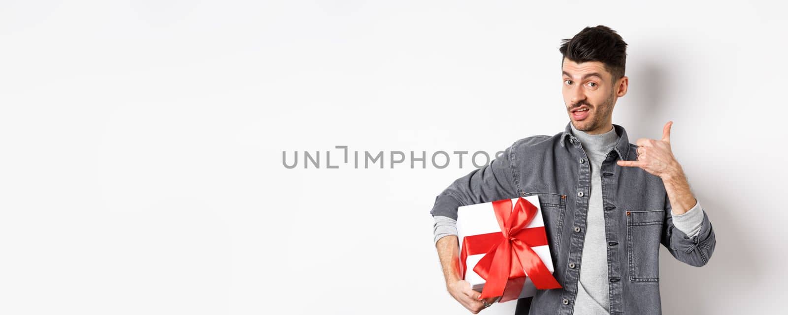 Macho guy with gift box show call me phone gesture, flirting with you, standing boastful on white background. Relationship and Valentines day concept by Benzoix