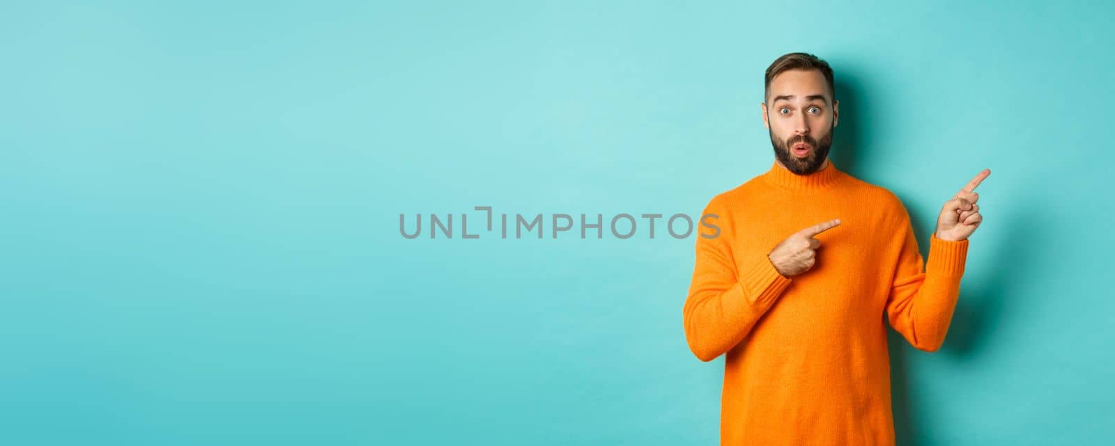 Amazed guy showing advertisement, pointing fingers right at banner, standing against turquoise background.