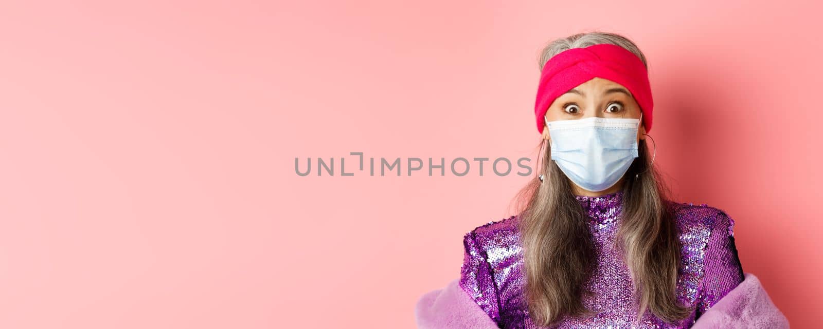 Covid-19, virus and social distancing concept. Close up of fashionable elderly woman in medical mask and headband staring surprised at camera, pink background.