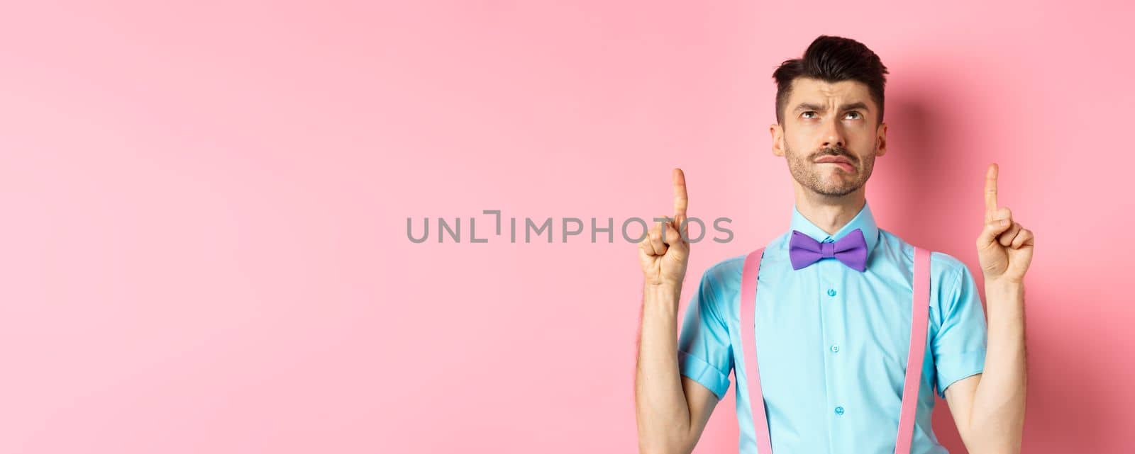 Jealous and sad funny man in bow-tie, biting lip and looking tempted at promo offer, pointing finger up, standing on pink background.