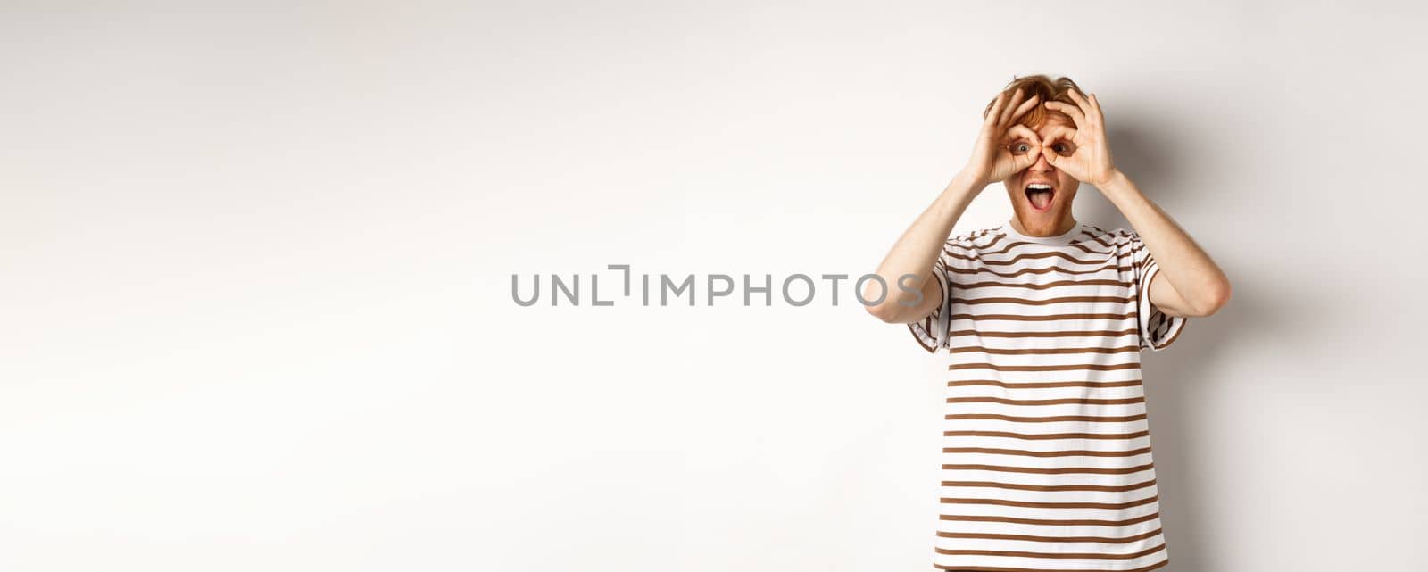 Impressed redhead man checking out promo offer, looking at camera from hand binoculars, smiling amazed, white background by Benzoix